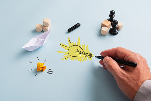 Hand of a businessman drawing light bulb with black marker on a blue background next to chess figures, origami boat and dice.