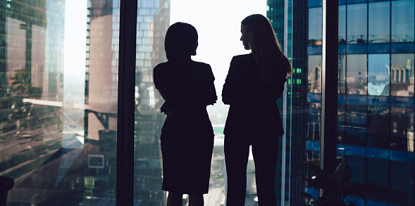 Back view of well dressed female colleagues in formal wear standing near window in contemporary workplace and enjoying cityscape while talking and looking at each other