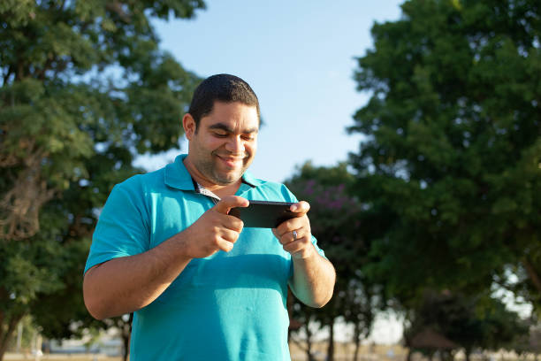 Portrait of a young Latin man in a natural park jaunt his cellphone at a park, surrounded by beautiful trees.