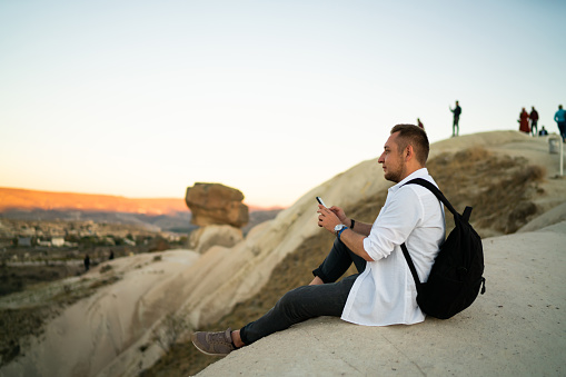 Side view of brown haired bearded male in white shirt with backpack using mobile phone watching natural landscape sitting on rock