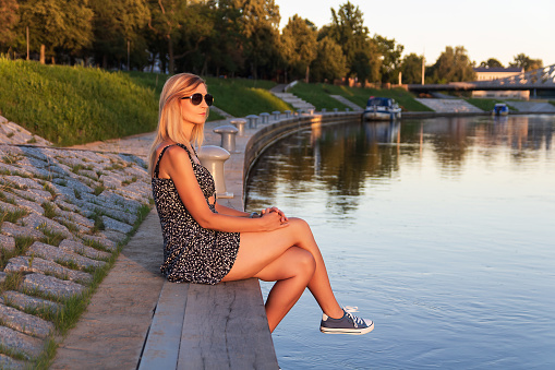 Portrait young blonde woman with sunglasses sitting in river Vltava port, city Ceske Budejovice at sunset