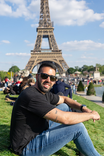 young man sitting on the lawn wearing sunglasses with the eiffel tower in the background, in Paris.