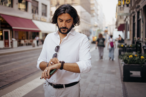 Serious handsome businessman talking on the cell phone while checking the time in modern city street. Busy male person arranging a business meeting.