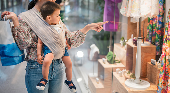 Asian woman window shopping while shopping for clothes in an unrecognizable shopping mall with her infant baby boy in a baby carrier. New mother lifestyle after having a baby