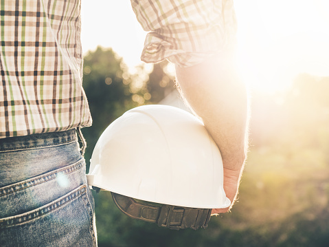Attractive man in work clothes, holding construction helmet in his hands against the background of trees, blue sky and sunset. Rear view, closeup, outdoors. Labor and employment concept