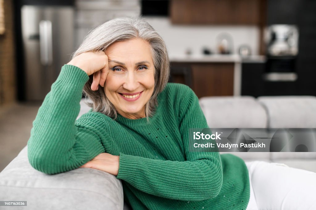 Beautiful senior woman at home Portrait of charming senior woman with grey hair, smiling mature female sits on the couch at ease and looks at the camera. Beautiful carefree middle-aged woman at home Mature Women Stock Photo