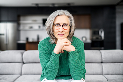 Senior mature gray-haired female freelancer grandparent portrait, businesswoman with hands on chin in glasses and green jumper sitting on couch in living room at home, confidently looking at camera
