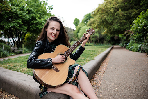 Portrait of smiling woman playing guitar in a park and looking at camera. She is wearing a dress and black leather jacket.