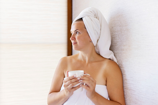 Beautiful young woman with a towel on her head holds a cup of coffee in the morning. Concept of domestic lifestyle.