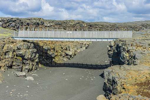 Lava rock landscape on Iceland Reykjanes peninsula. Metal bridge over the North American and Eurasian rifts. Lava stone wall with black sand and green grass in sunshine and clouds