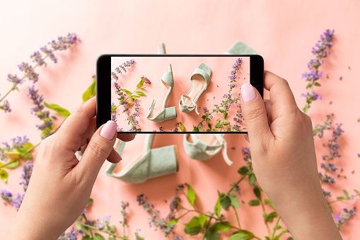 Woman taking photo of pastel mint green sandals shoes and meadow flowers with smartphone. Blogger, influencer or stylist capturing fashion footwear for social media. Pink background.