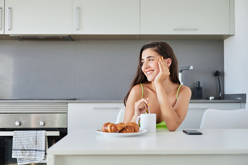 Young woman having breakfast in a modern kitchen at home. She has just got up in the morning. She is drinking coffee and eating a croissant.