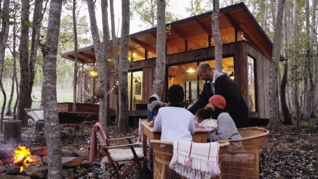 Family at table by campfire outside cabin in woods