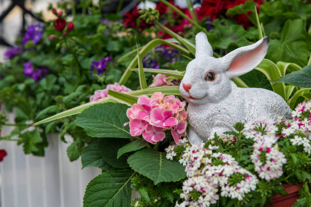 A figurine of a white rabbit stands in a flower bed near bright colorful geranium flowers. Copy space for your text. Easter decoration theme.