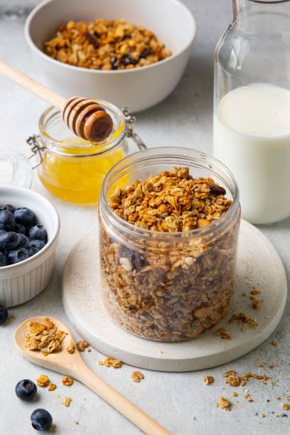 gesunde frühstückszutaten. hausgemachtes müsli mit rosinen, sonnenblumenkernen, haselnuss und erdnuss im offenen glas, milch- oder joghurtflasche, honig und blaubeeren auf grauem hintergrund. selektiver fokus. - jar oatmeal granola glass stock-fotos und bilder