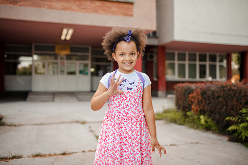 Little girl standing in front of her school on her first school day and waving towards camera
