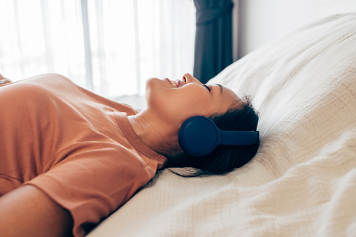 Young Asian woman, lying on the bed, listening to music on her headphones with her eyes closed.