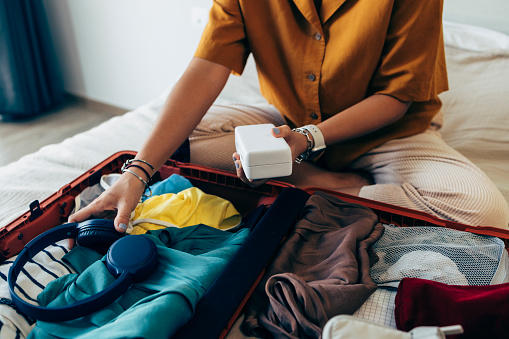 Anonymous woman sitting on the bed in a hotel room, packing her clothes in a suitcase.