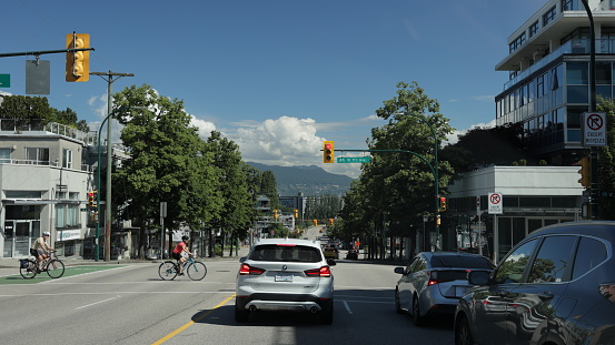 Vancouver, Canada - July  1, 2022: Cyclists ride east along the West 7th Avenue bicycle route. View facing north on Burrard Street in the Kitsilano neighborhood, a popular commerical and residential district. Background shows the Coast Mountain Range under summer clouds.