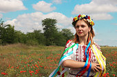 Portrait of Ukrainian young girl in national costume with wreath on her head on background of field with poppies and blue sky . Ethnic Ukrainian costume. Patriotic themes