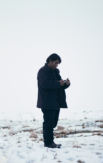Side view of native American male in outerwear standing in snowy meadow in winter and surfing mobile phone