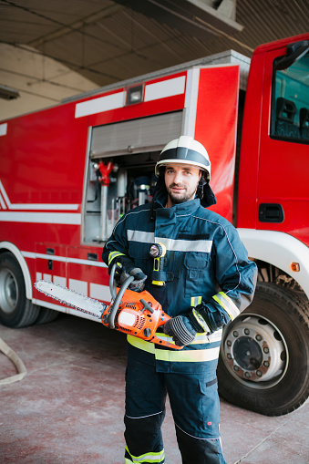 Portrait of a professional fireman standing in a fire station garage next to the fire truck, holding a chainsaw in his hands and looking at the camera
