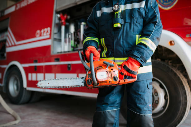 Unrecognizable professional fireman standing in a fire station garage next to the fire truck, holding a chainsaw in his hands