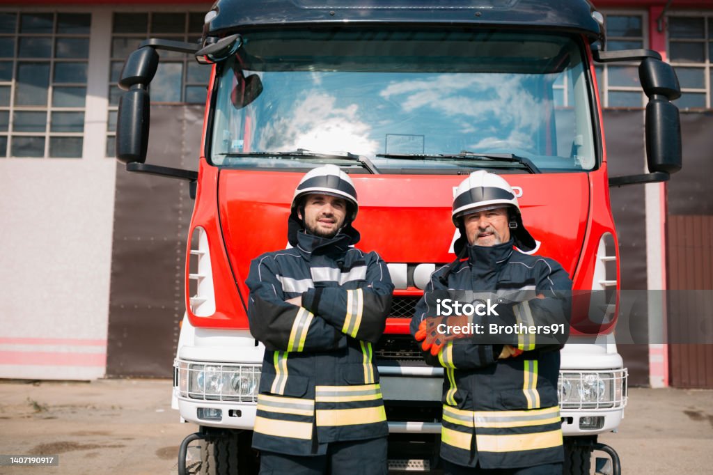 Two firefighters in the fire station Portrait of two firefighters standing in front of the fire truck, dressed in their uniform and having their arms crossed, looking at the camera Fire Station Stock Photo