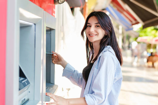Cute brunette woman wearing shirt standing on city park, outdoors standing in front of ATM machine, smiling and holding credit or debit card. Looking at the camera with smiles. stock photo