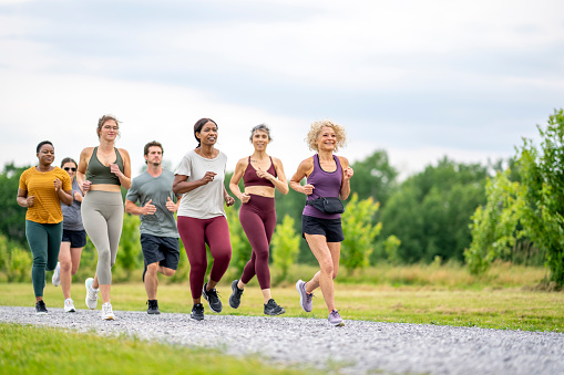 A large group of mature adults are seen running together as part of a club.  They are dressed comfortably in athletic wear as they run down the road on a warm summer day.
