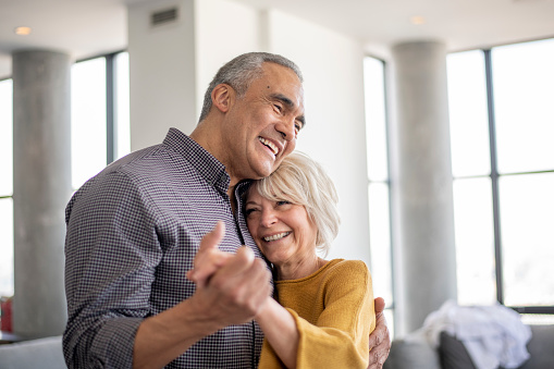 A senior couple hold hands as they dance around the living room together while at home.  They are both dressed casually and smiling as they enjoy the time to connect and bond together.