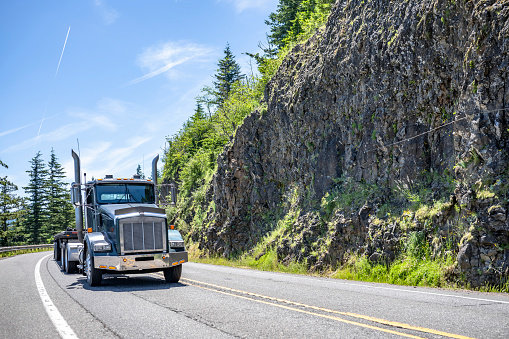 Powerful black bonnet day cab classic big rig semi truck tractor transporting cargo on flat bed semi trailer driving on the narrow winding mountain highway road with rock cliff in Columbia Gorge area