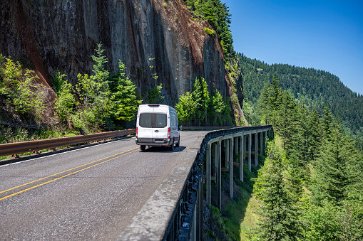 Popular commercial freight compact white cargo mini van for deliveries and small business running on the overpass mountain road with rock wall on the one side and abyss on the another side