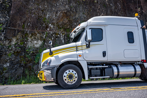 Low cab profile to improve aerodynamics big rig white semi truck tractor with oversize load sign on the bumper and flashing lights on the roof running on the mountain road with cliff on the side