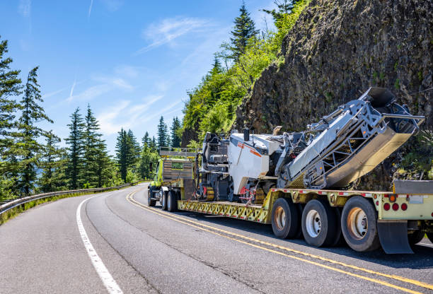 istock Light green day cab big rig semi truck transporting construction equipment on step down semi trailer running on the winding mountain highway road 1407216429