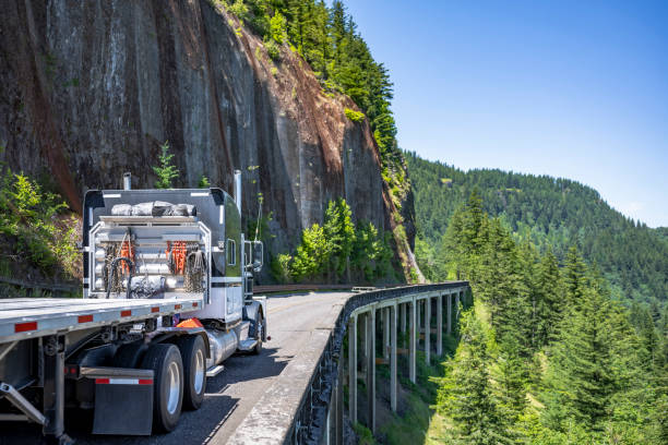 istock Pro classic black big rig semi truck with empty flat bed semi trailer going to warehouse for the next load running on the bridge around the rock cliff on mountain road in Columbia Gorge 1407216645