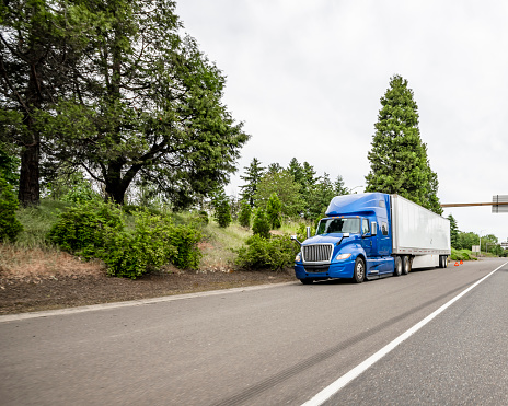 Industrial freight broken blue big rig semi truck with dry van semi trailer standing out of service on the road shoulder waiting for mobile road assistant or towing truck for tow to repair shop