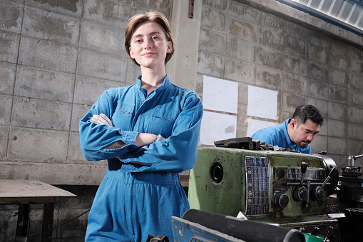 Portrait of Young White female industry worker stands, arms crossed, looks at camera and smiles, male partner works with machine behind her in manufacturing factory, professional mechanical engineers.