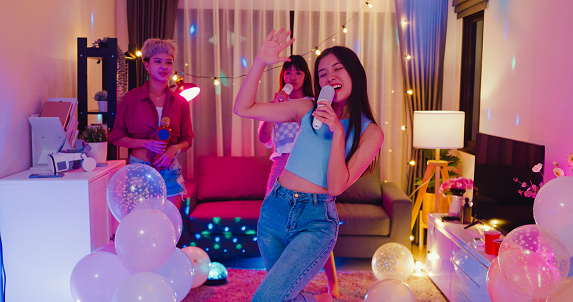 Young Asian female friends singing karaoke having fun at colorful house party at night. Lifestyle together concept.
