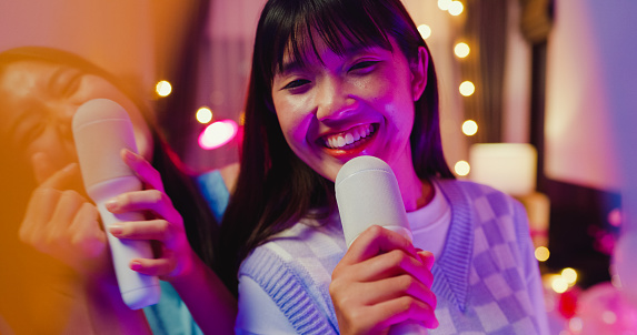Close up of young Asian female friends singing karaoke having fun at colorful house party at night. Lifestyle together concept.