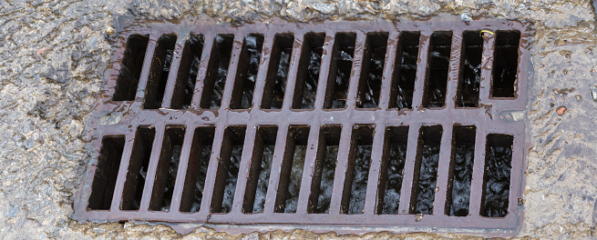 Cast iron grid of the storm drain mounted in a concrete surface with flowing down rainwater and small debris during a rain, panoramic view close-up