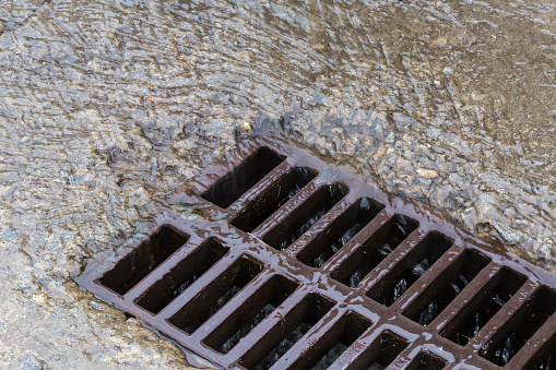 Fragment of the cast iron grid of the storm drain mounted in a concrete surface and flowing down rainwater with small debris during a rain