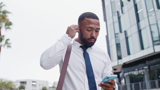 Businessman texting on cellphone, networking with clients on the go. Black professional looking contemplative, listening to music on wireless earphones. Corporate worker using phone in morning commute
