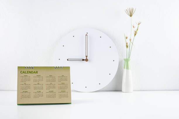 Clear, clean theme white office desk concept : Business working space and office supply on a table e.g flipped desktop calendar, analog clock, two toned vase near a white wall.