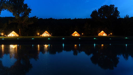 Tents with illumination in front of a lake at glamping, forest around, night