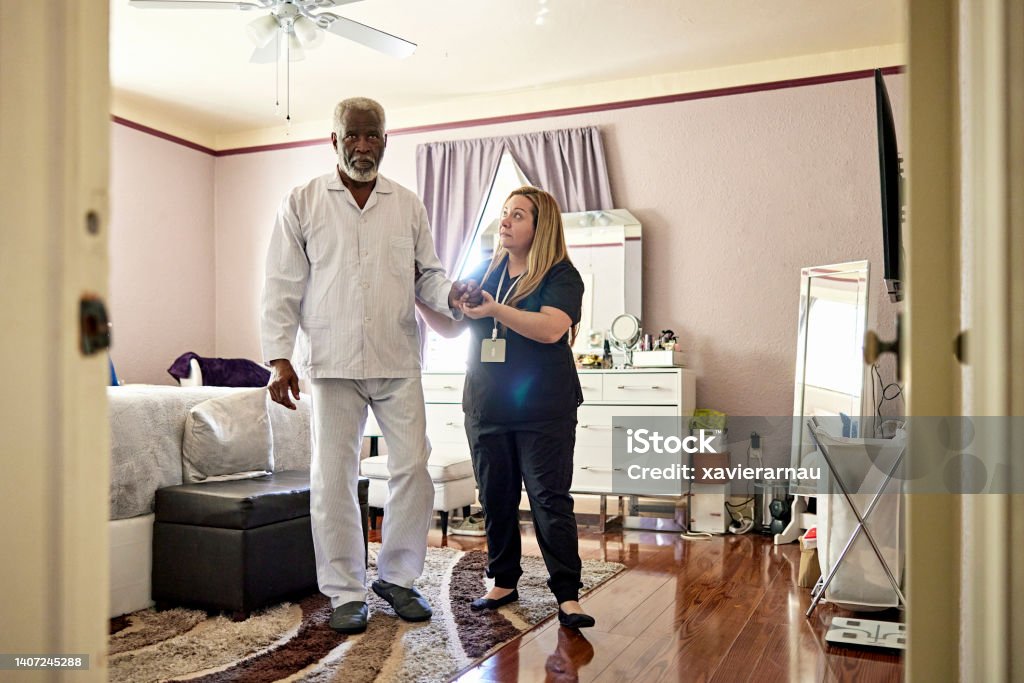 Caregiver helping senior man take first steps in the morning Full length front view of early 70s Black man in pajamas and slippers walking in bedroom with assistance of Latin American woman in uniform. Home Caregiver Stock Photo