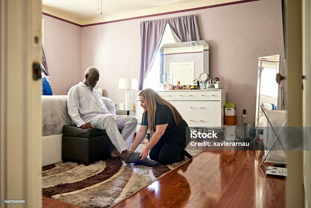 Early 70s Black man receiving assistance with his slippers Full length view from bedroom doorway as Latin American home caregiver helps senior client in pajamas get up and start his day. Community Outreach Stock Photo