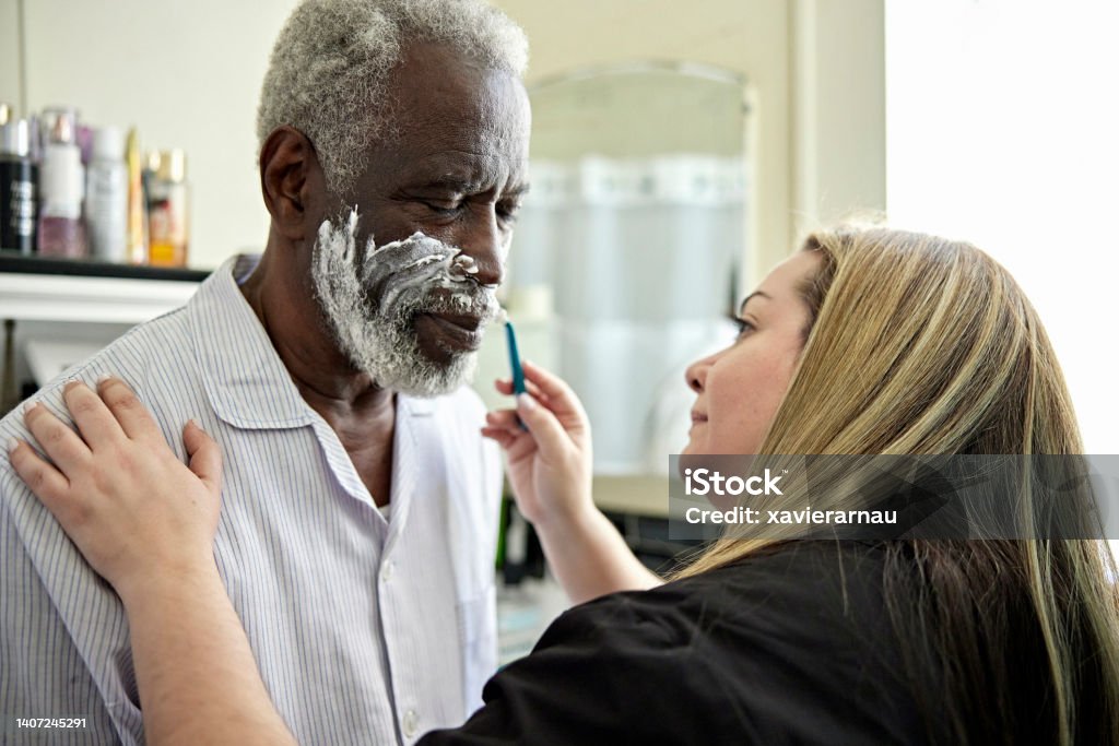 Latin American caregiver shaving senior Black client Head and shoulders view of mid adult woman in uniform assisting early 70s man in pajamas with his morning routine. Senior Adult Stock Photo