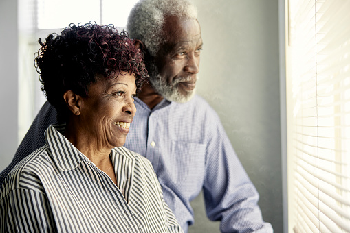 Three-quarter front view of bearded man and smiling woman, both in their 70s and wearing smart casual attire, looking outside with expressions of wellbeing.