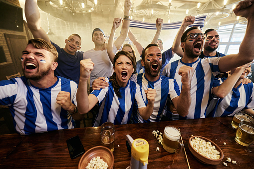 Large group of cheerful sports fans screaming while celebrating the success of their team during a game on TV in a bar.